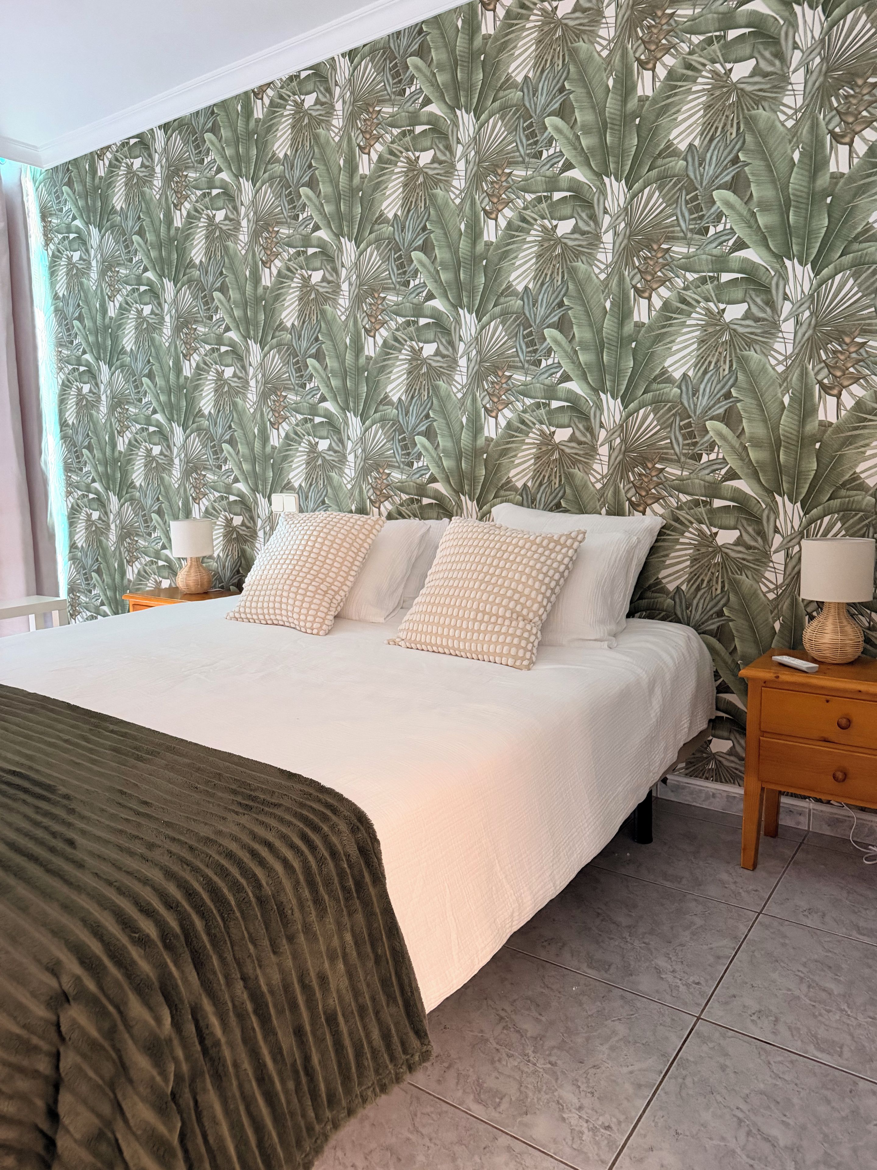 Un dormitorio con una cama grande, una pared con estampado de hojas tropicales, una mesita de noche con lámpara y un cajón de madera.