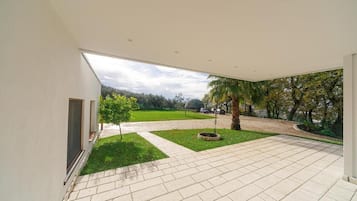A modern building with a spacious patio area, a small lawn, and a view of a park with trees and a pathway.