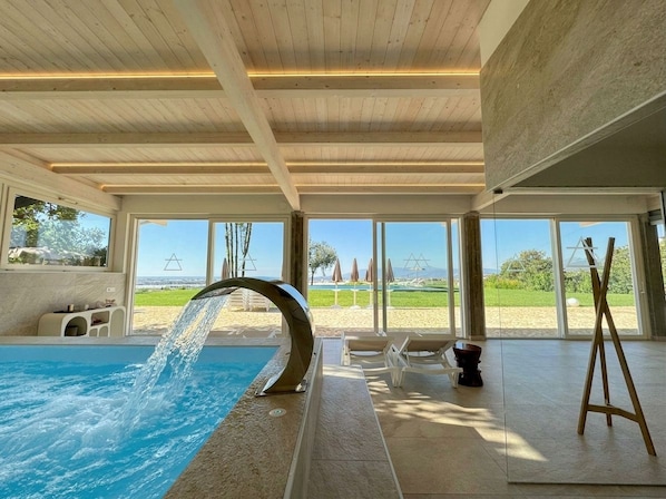 An indoor swimming pool with a water feature, large windows, and a view of a beach.