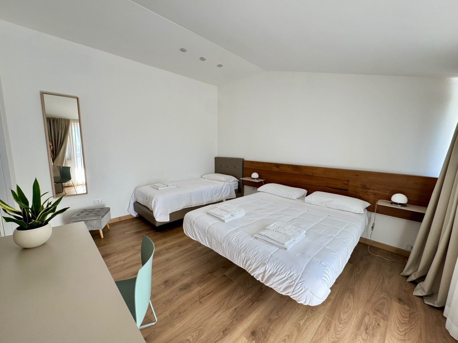 A hotel room with two beds, a desk, a chair, a potted plant, and a mirror.