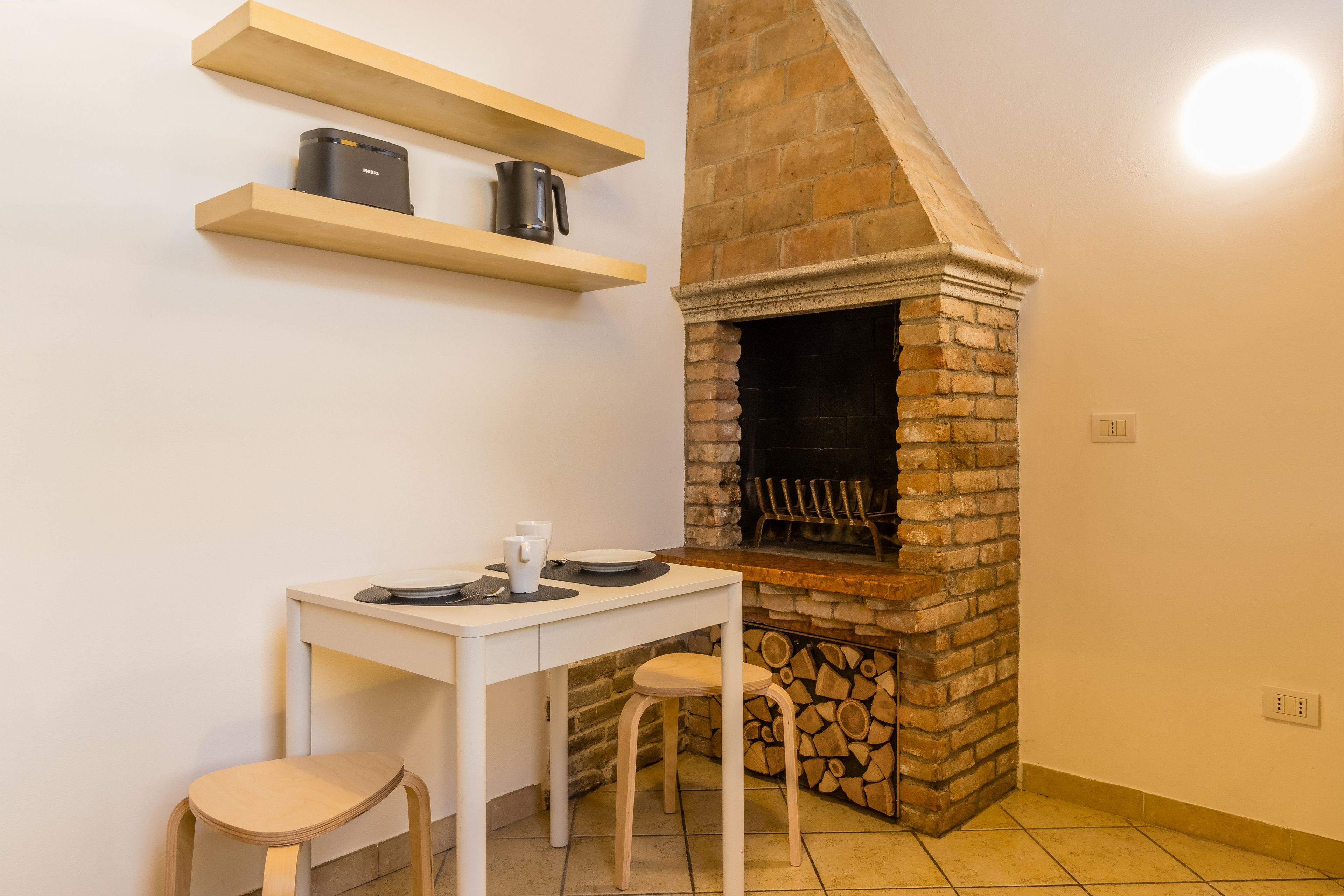 A kitchen with a brick fireplace, a white table with stools, and a shelf above the counter.