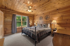 A wooden-paneled room with a bed, two bedside lamps, a window with curtains, and a ceiling fan. - Cubby Bear Cabin • Cabin with Mountain Views • Pool Table & Arcade (Sevierville)