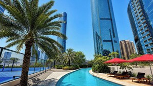A swimming pool with lounge chairs and umbrellas, surrounded by palm trees and modern buildings. - Vanilla Homes Premium 1BR in Gate Tower (Abu Dhabi)