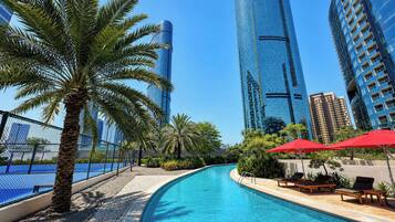 Una piscina con sillones de descanso y sombrillas, rodeada de palmeras y edificios modernos.