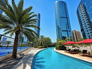 Una piscina con sillones de descanso y sombrillas, rodeada de palmeras y edificios modernos.