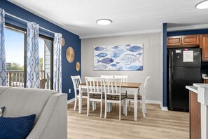 A dining area with a wooden table and white chairs, a blue wall with a fish-themed artwork, and a kitchen with a black refrigerator. - B126 at The Myrtle Beach Resort (Myrtle Beach)