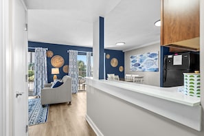 Eine moderne Küche mit einem schwarzen Kühlschrank, ein Essbereich mit weißen Stühlen und ein Wohnzimmer mit einer Couch und gemusterten Vorhängen.