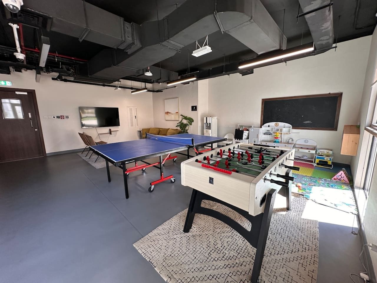 Een kamer met een tafeltennistafel, een tafelvoetbaltafel en een groot raam.