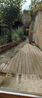 Una terraza de madera que conduce a un muro de piedra con una ventana, rodeada de follaje y un muro de ladrillo.