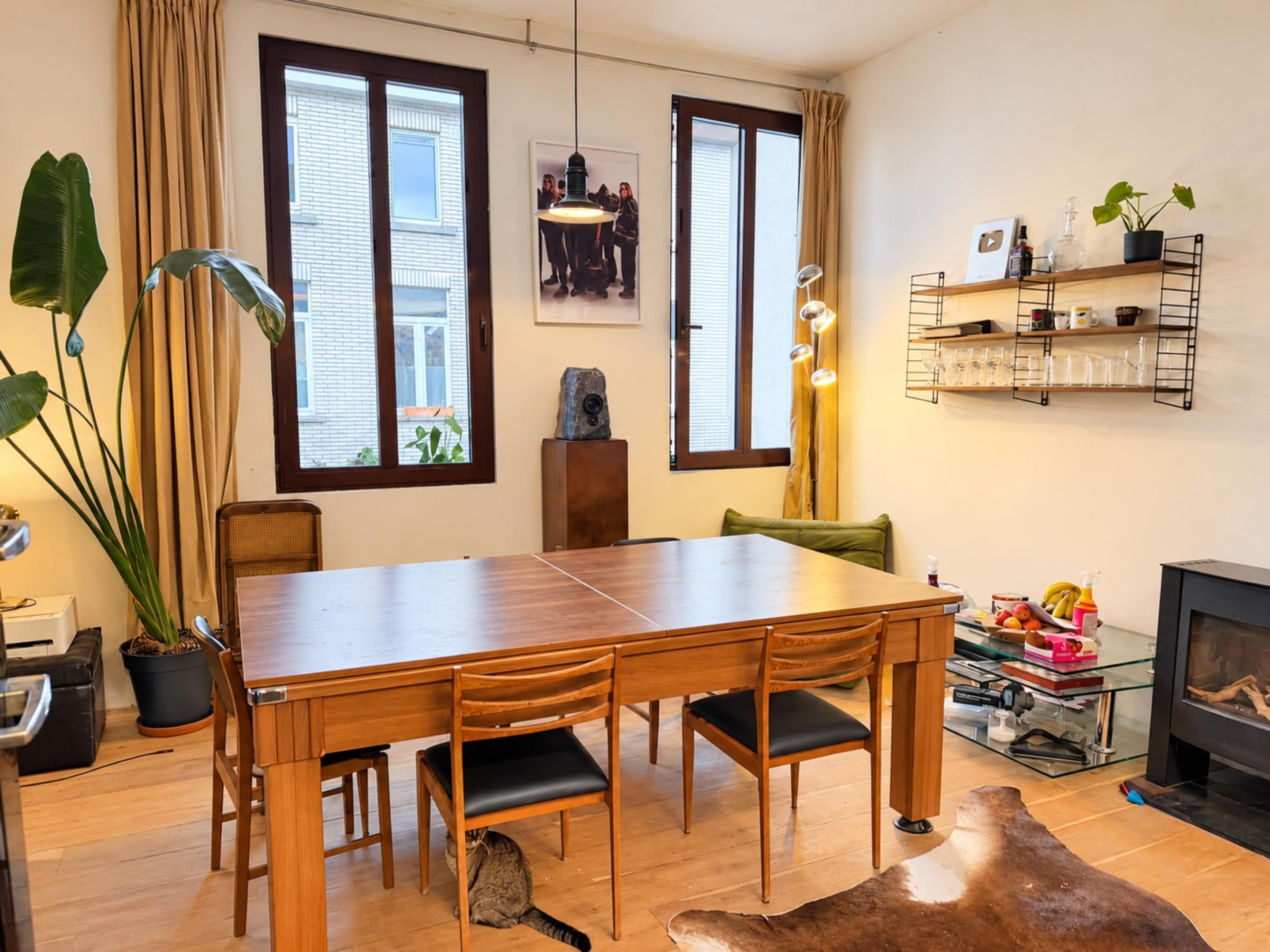 Een eetgedeelte met een houten tafel en stoelen, een open haard en uitzicht op het omliggende landschap door grote ramen.