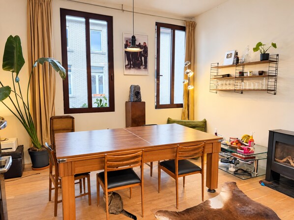 A dining area with a wooden table and chairs, a fireplace, and a view of the outside through large windows.