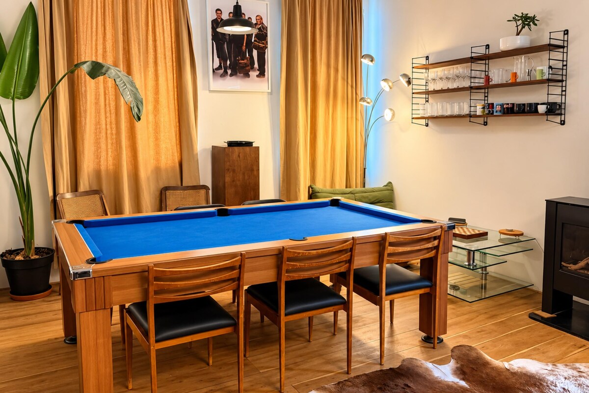 A pool table with chairs, a glass coffee table, a sofa, a fireplace, and a shelf with glassware.