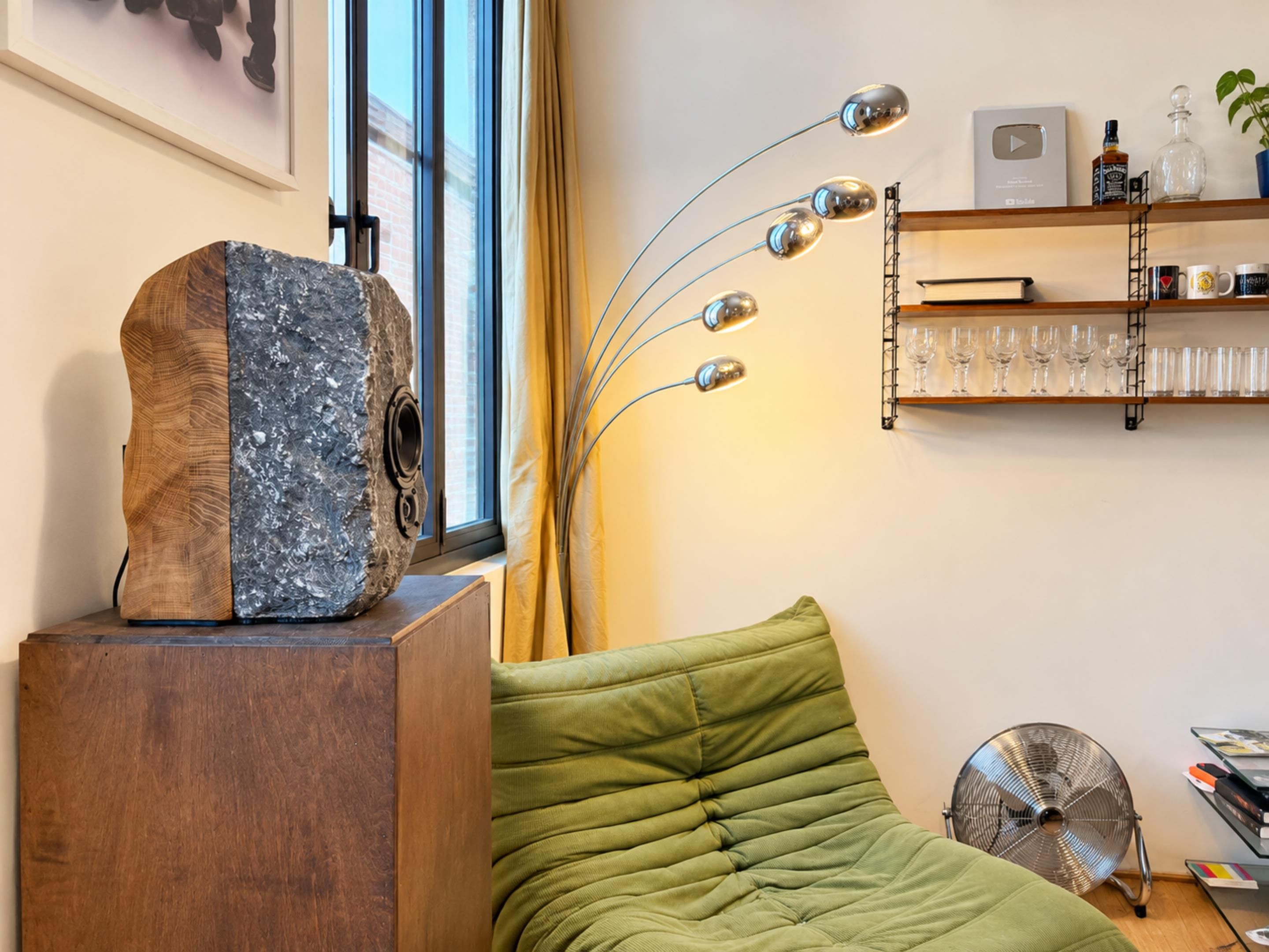 Een kamer met een houten beeld op een voetstuk, een groene gewatteerde bank, een vloerventilator en een plank met boeken en glazen.