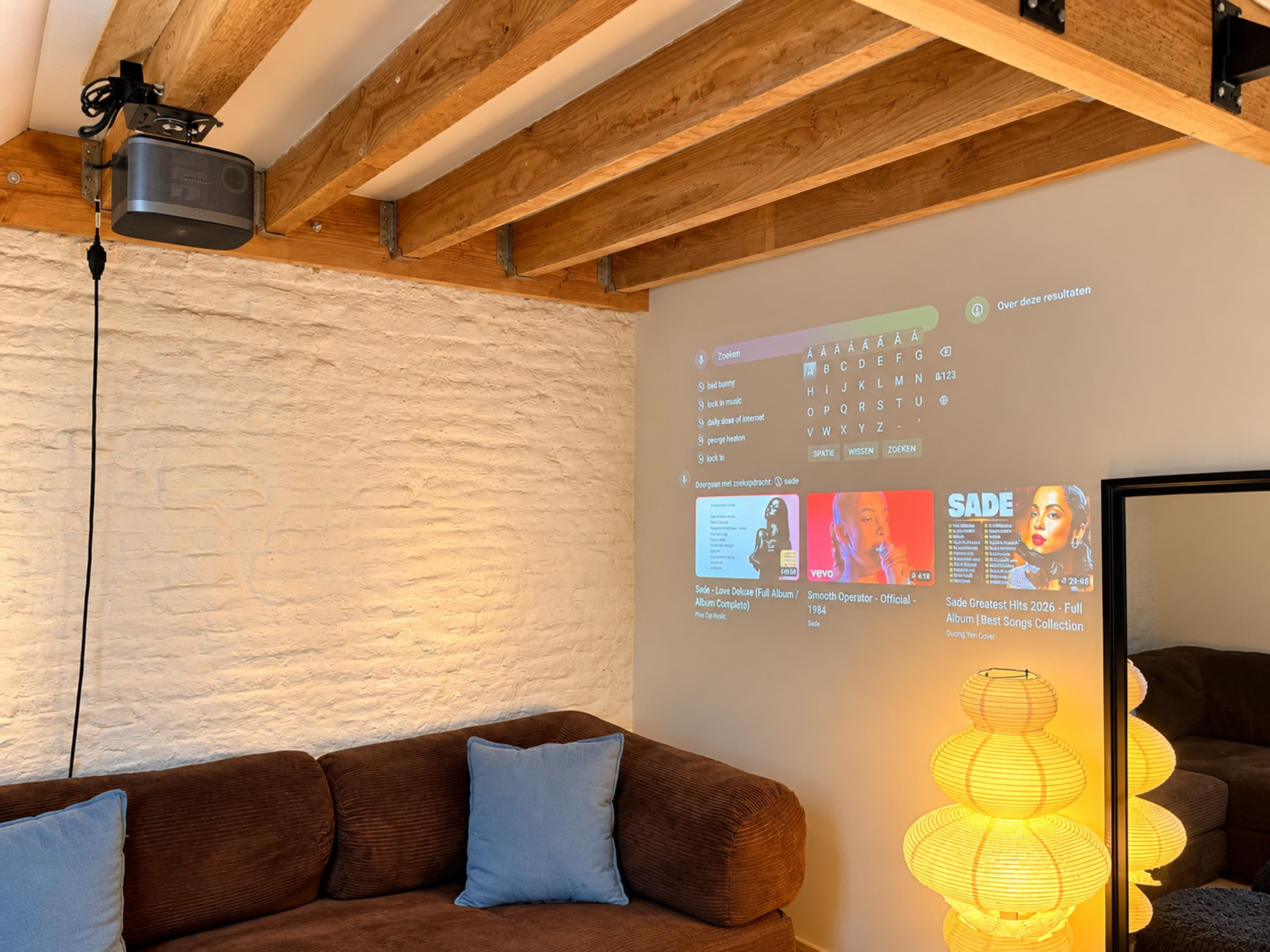 Quarto com teto de madeira, projetor exibindo capas de álbuns musicais, sofá marrom e luminária fixada na parede.