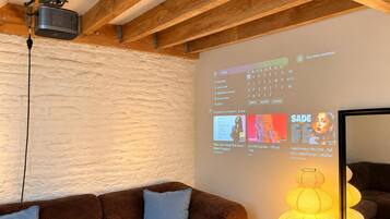 Quarto com teto de madeira, projetor exibindo capas de álbuns musicais, sofá marrom e luminária fixada na parede.