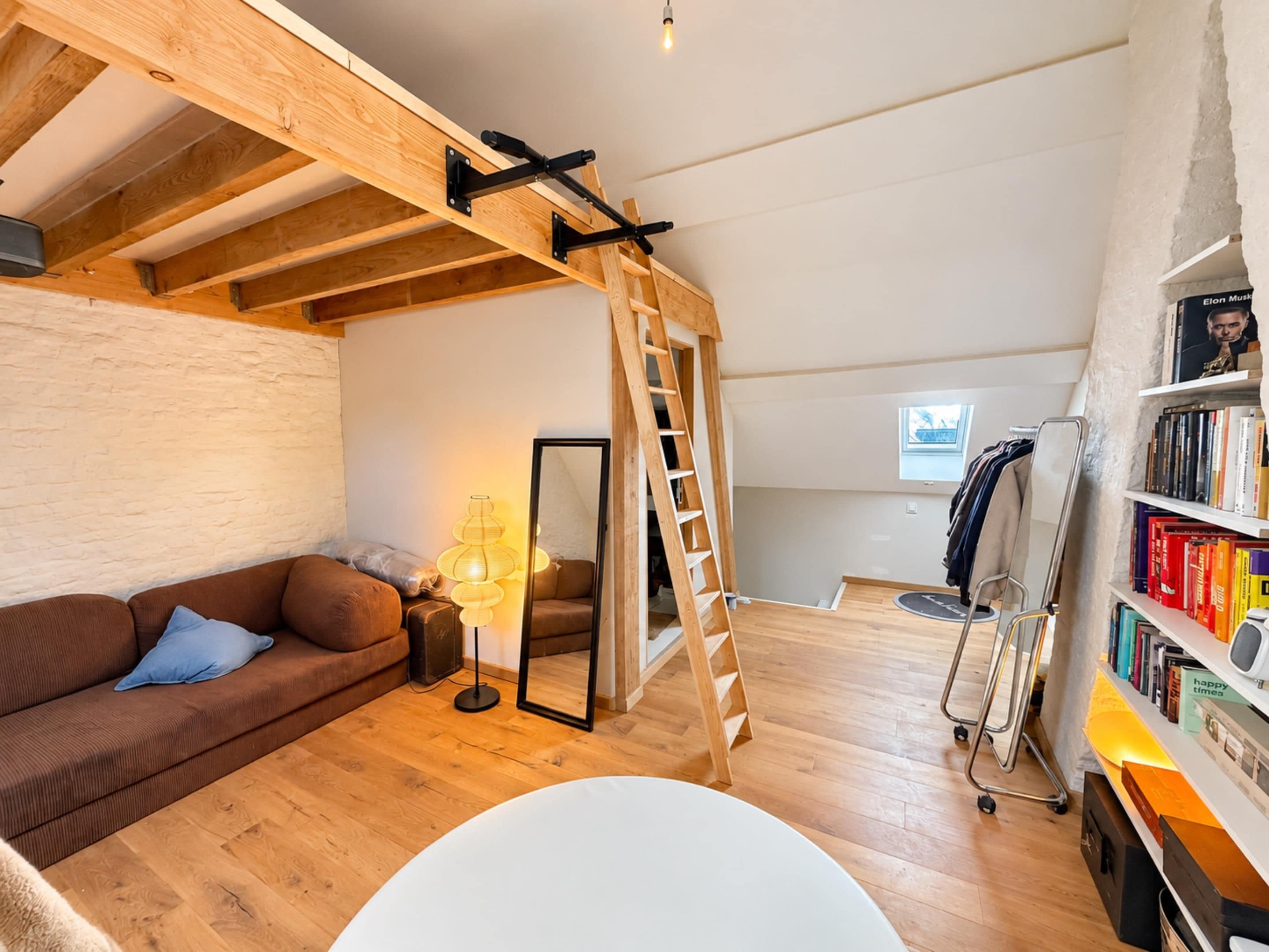 Een kamer in loftstijl met zichtbare houten balken, een bruine bank, een staande lamp, een ladder en een boekenkast.