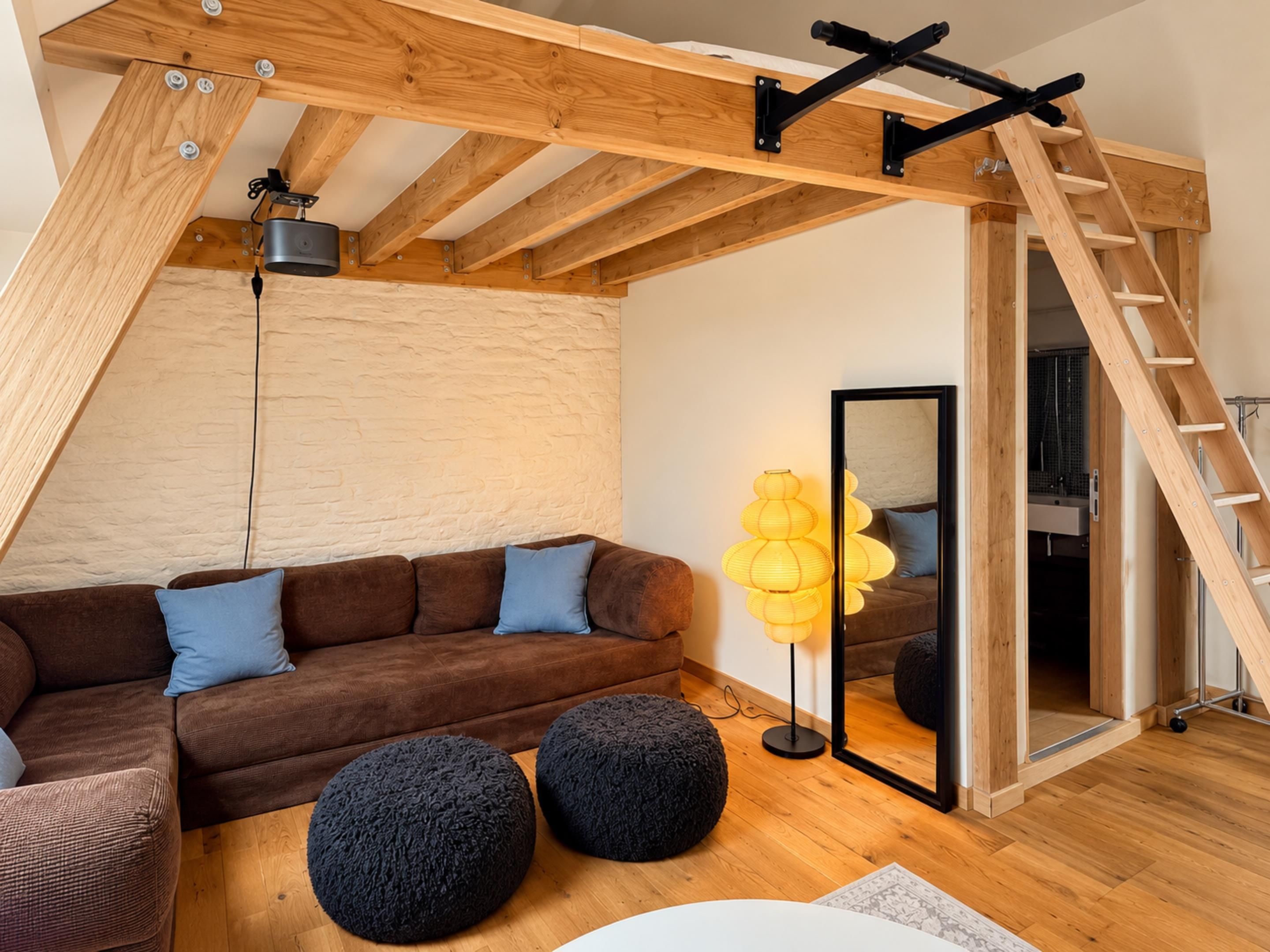 Een loft-achtige kamer met een houten plafond, een bruine hoekbank, twee zwarte poefs, een vloerlamp en een ladder naar een hoger niveau.