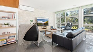 Ein modernes Wohnzimmer mit Flachbildfernseher, Ledermöbeln, einem Bücherregal und großen Fenstern mit Blick ins Freie.