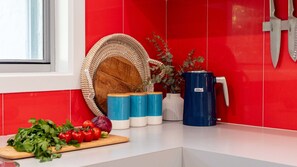 Eine Küchenarbeitsplatte mit Schneidebrett, frischen Tomaten und Kräutern. Blaue und weiße Behälter, ein blauer Wasserspender und ein gewebter Korb sind vorhanden.