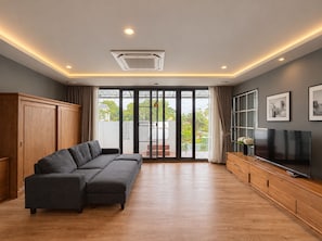 A modern living room with a sectional sofa, a flat-screen TV, and a sliding glass door leading to a balcony. - Yun Prestige Villa: 6 Bedrooms, Private Pool & Garden (Tambon Nong Phueng)