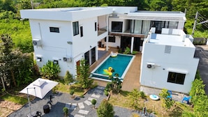 Uma casa moderna de dois andares com piscina, cercada por vegetação e um espaço de lazer com piso pavimentado.