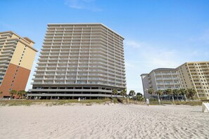 A multi-story hotel with a flat roof and balconies on each floor, located on a sandy beach with palm trees. - Seawind 1109 by Liquid Life (Gulf Shores)