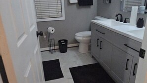 A bathroom with a toilet, sink, and mirror. There is a black rug on the floor and a window with blinds. - Charming 3-bedroom house in Rex near Atlanta (Rex)