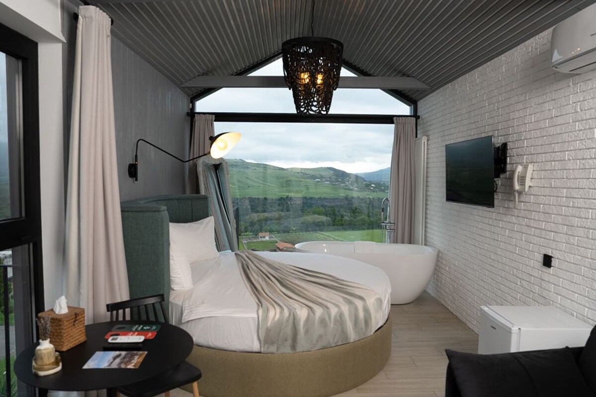 A modern hotel room with a circular bed, a small table with a book, a chair, a flat-screen TV, and a large window with a view of the countryside.