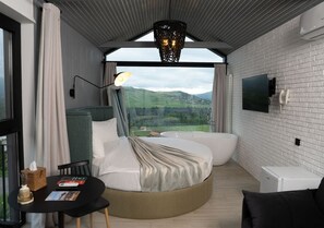 A modern hotel room with a circular bed, a small table with a book, a chair, a flat-screen TV, and a large window with a view of the countryside. - Shakaryan Rancho&Restaurant (Dsegh)