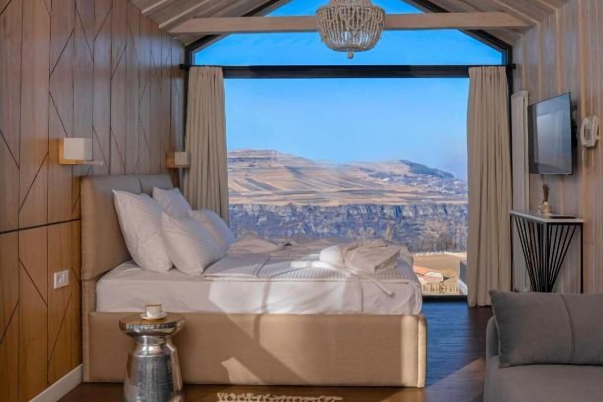 A bedroom with a large bed, a view of the landscape, a chandelier, and a television.