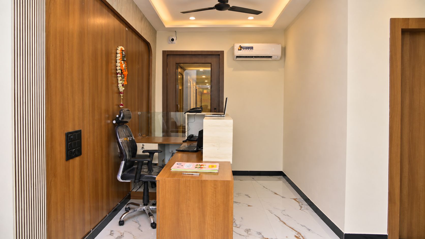 A modern office space with a wooden reception desk, a black office chair, a ceiling fan, and an air conditioning unit.