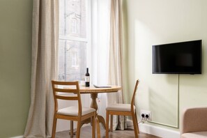 A room with a dining table, two chairs, a bottle of wine, a television on the wall, and a window with curtains. - Bright 1 Bed Flat | Central Edinburgh, Sleeps 2 (Edinburgh)