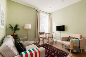 A living room with a sofa, a television, a dining table with chairs, a lamp, and a window with curtains. - Bright 1 Bed Flat | Central Edinburgh, Sleeps 2 (Edinburgh)