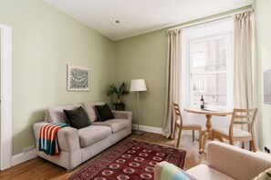 A living room with a sofa, a coffee table, a lamp, and a view of the street through a window. - Bright 1 Bed Flat | Central Edinburgh, Sleeps 2 (Edinburgh)