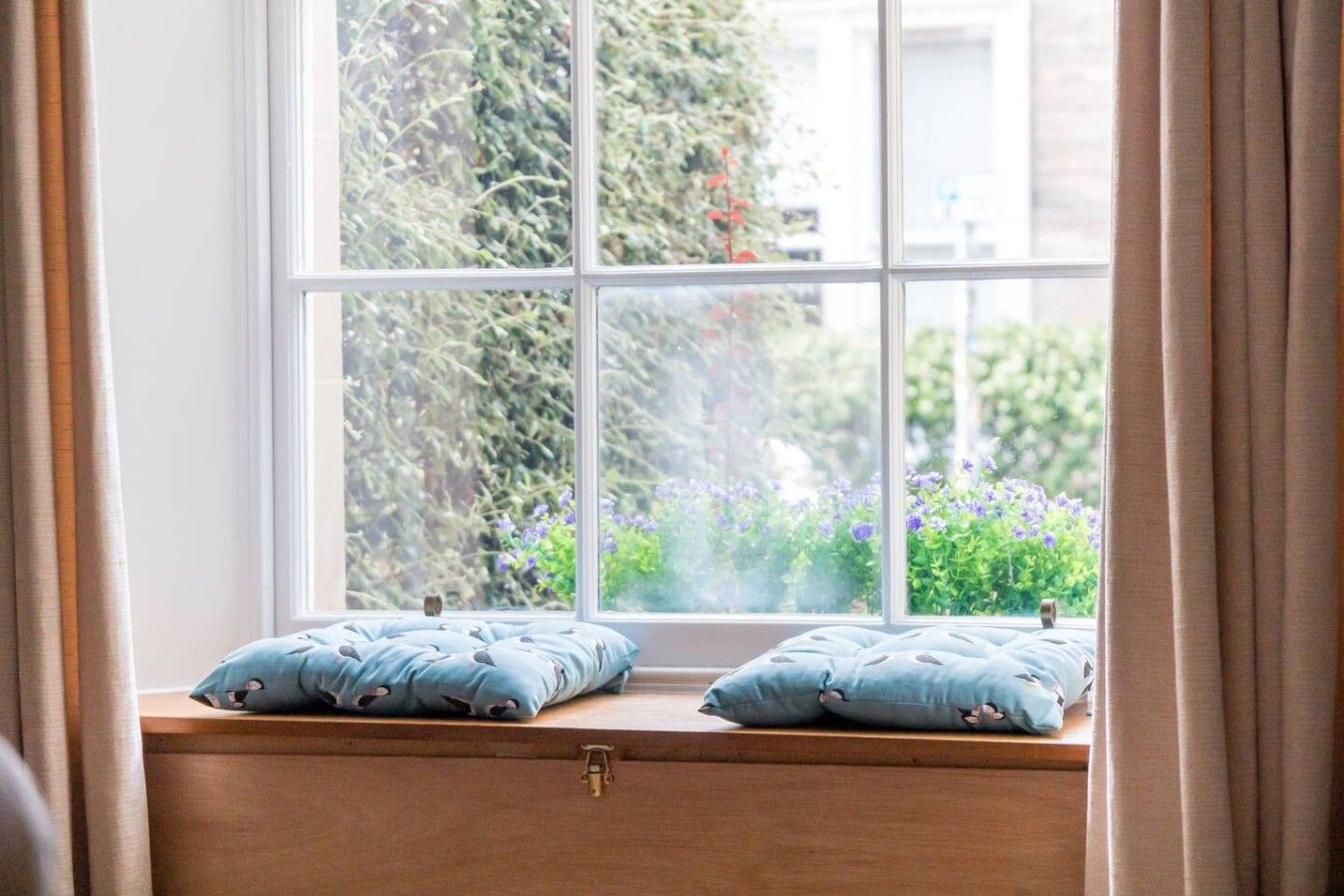A window with a view of greenery and flowers, a wooden bench with cushions, and curtains.