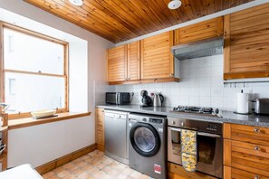A kitchen with wooden cabinets, a washing machine, a microwave, a stove, and a window with a bowl on the counter. - Potter`s House | Central 2 Bed, Sleeps 4 (Edinburgh)