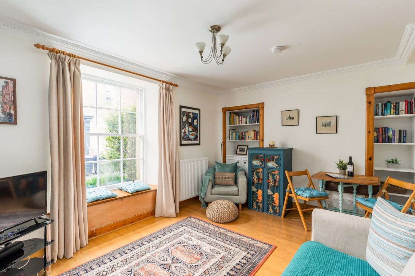 A cozy living room with a large window, a bookshelf, a TV, and a patterned rug.