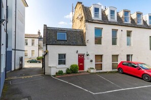 A residential street with a red car parked in a driveway, a white building with a red door, and a black car in the background. - Potter`s House | Central 2 Bed, Sleeps 4 (Edinburgh)