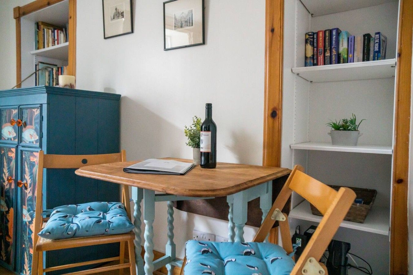 A room with a wooden table, two chairs, a cabinet, and shelves with books and a plant.