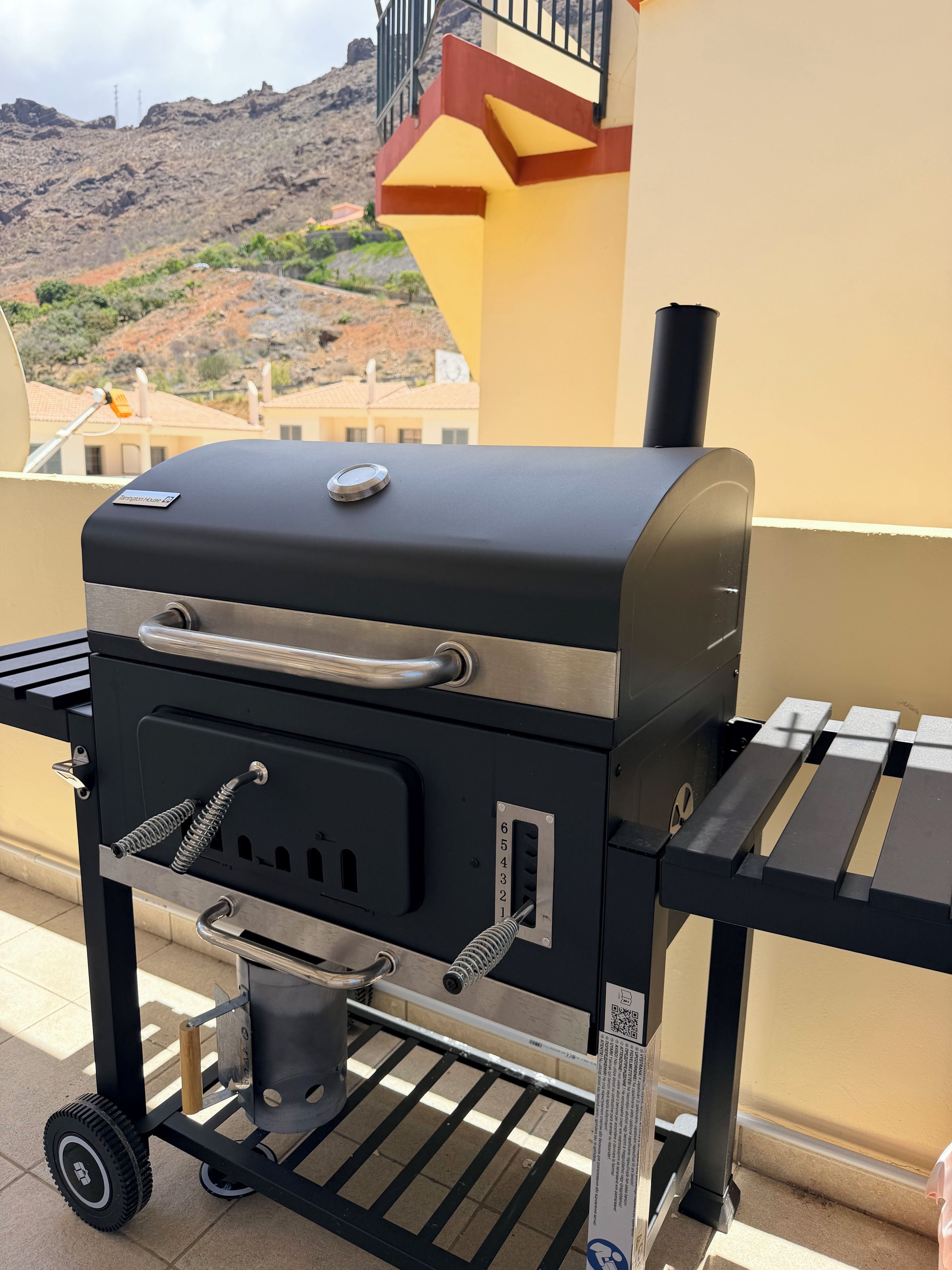 A portable barbecue grill with wheels, a digital thermometer, and a control panel, set against a building with a yellow facade and a mountainous backdrop.