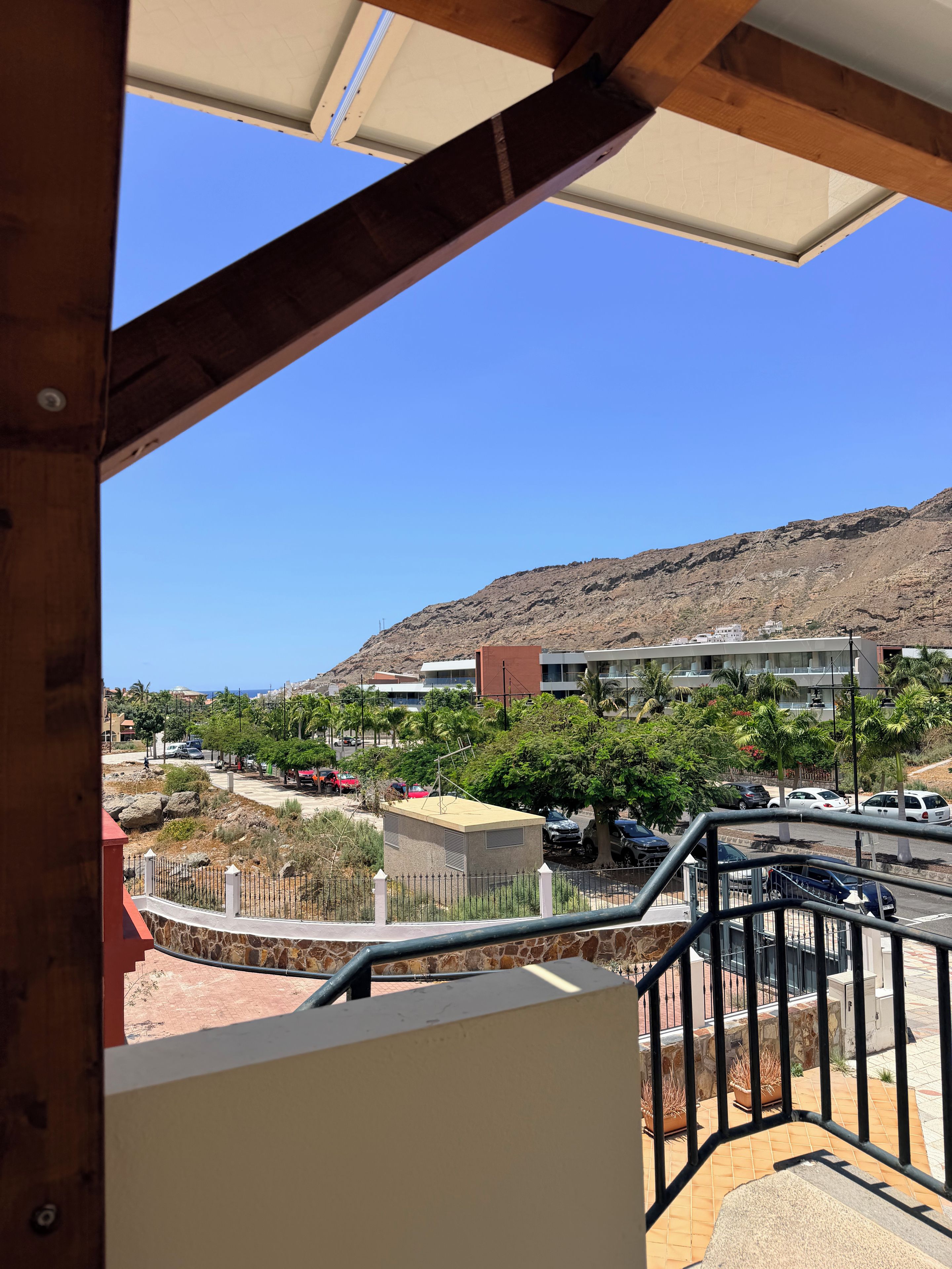 Vista desde un balcón sobre un estacionamiento con coches, un edificio de techo plano y una montaña al fondo.
