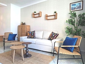 Un saló amb un sofà, dues butaques, una taula de centre i una catifa. Hi ha dues baldas penjades a la paret amb plantes, una planta gran en test i un quadre amb marc.