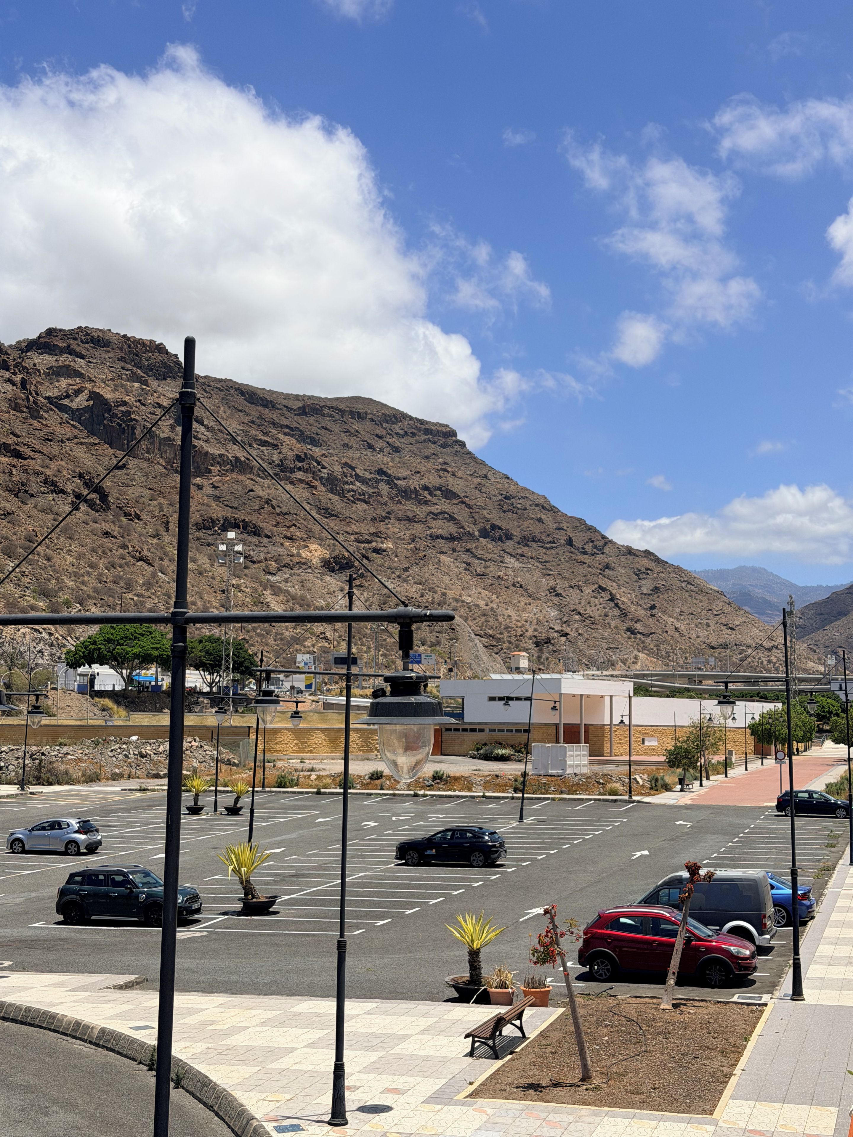 Un estacionamiento con autos, un banco y un edificio con una montaña al fondo.