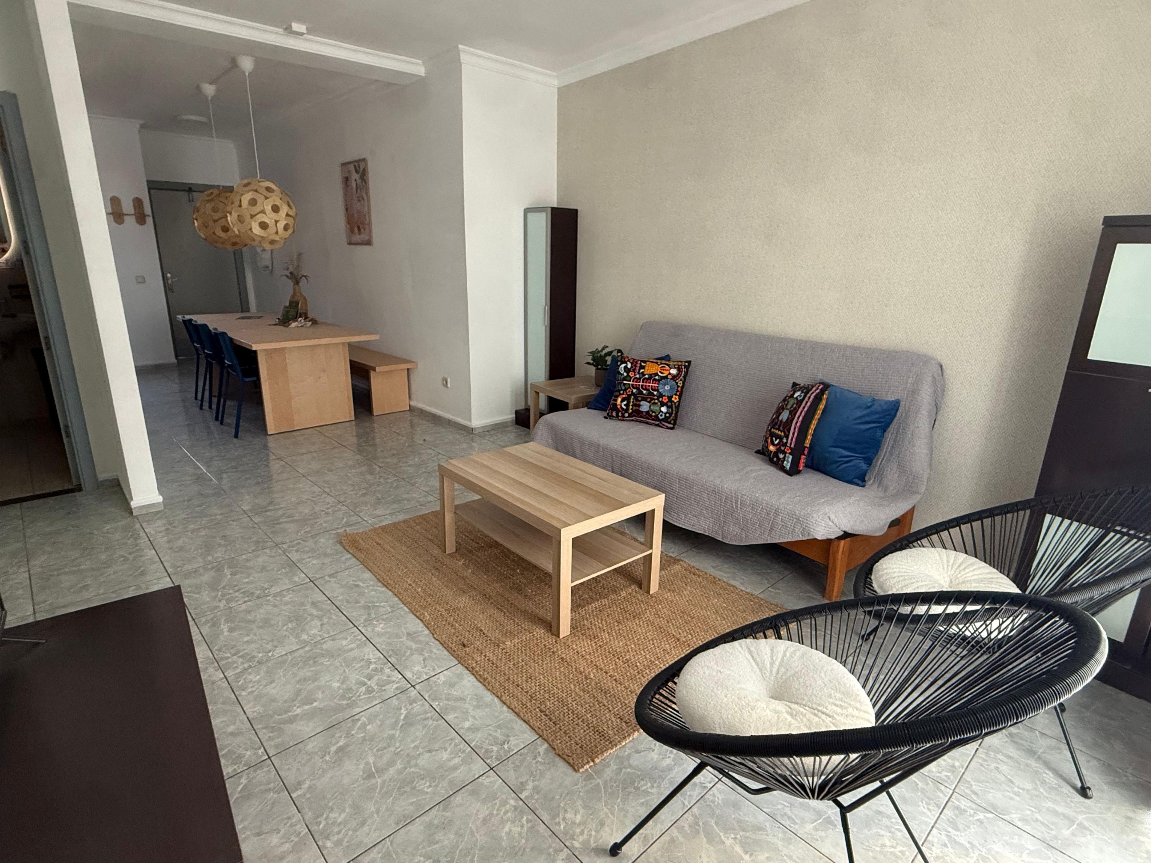 A modern living room with a sofa, a coffee table, a dining area with chairs, and a black chair with a cushion.