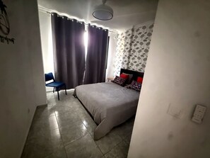 Un dormitorio con cama, una silla, un ventanal con cortinas y un papel tapiz estampado en la pared.