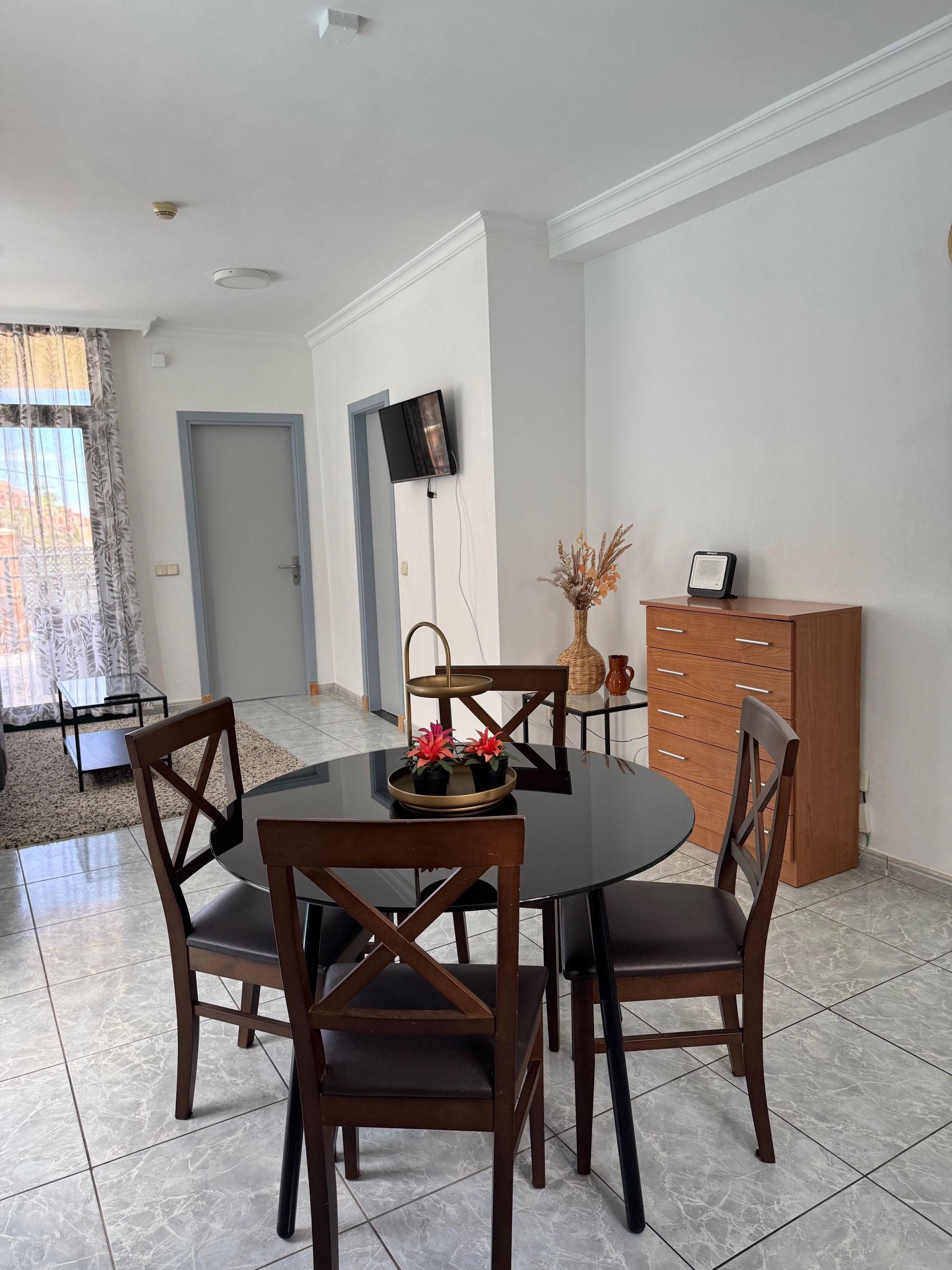 Comedor con una mesa redonda, cuatro sillas de madera, una mesita auxiliar con un florero y un televisor montado en la pared.