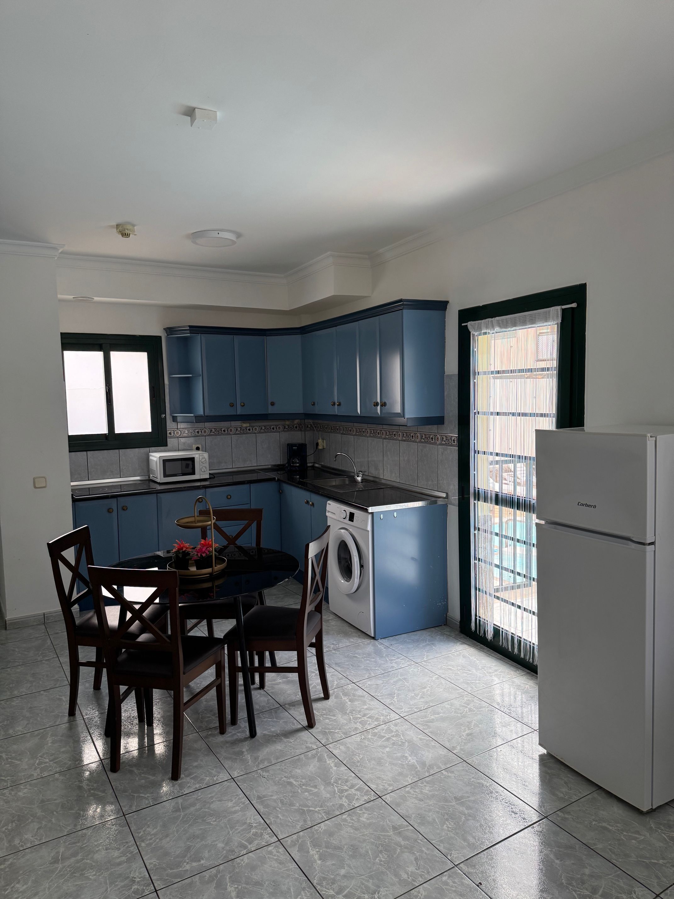 Cocina compacta con armarios azules, lavadora, microondas y un comedor con sillas y mesa.