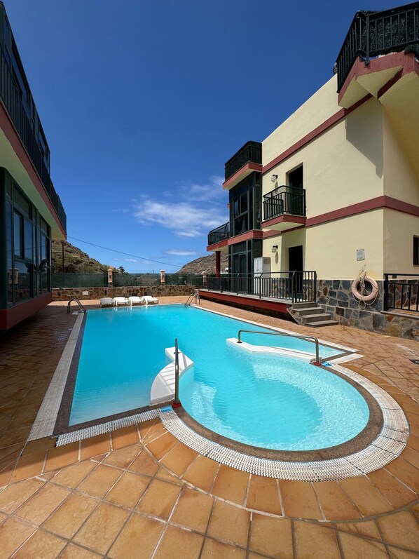 Una piscina con borde de baldosas, rodeada de edificios con balcones y un cielo despejado.