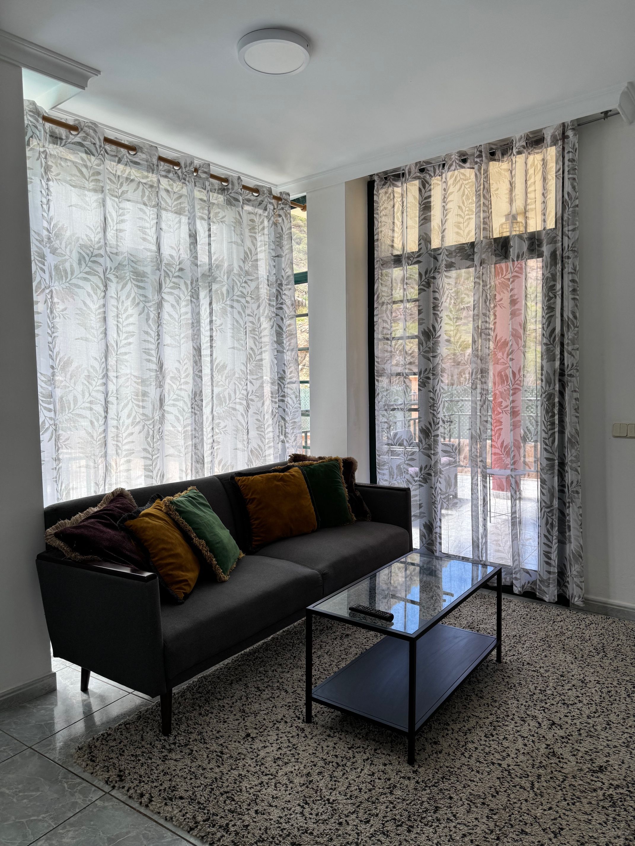 Un salón moderno con un sofá, una mesa de centro de vidrio y vistas a través de cortinas traslúcidas.