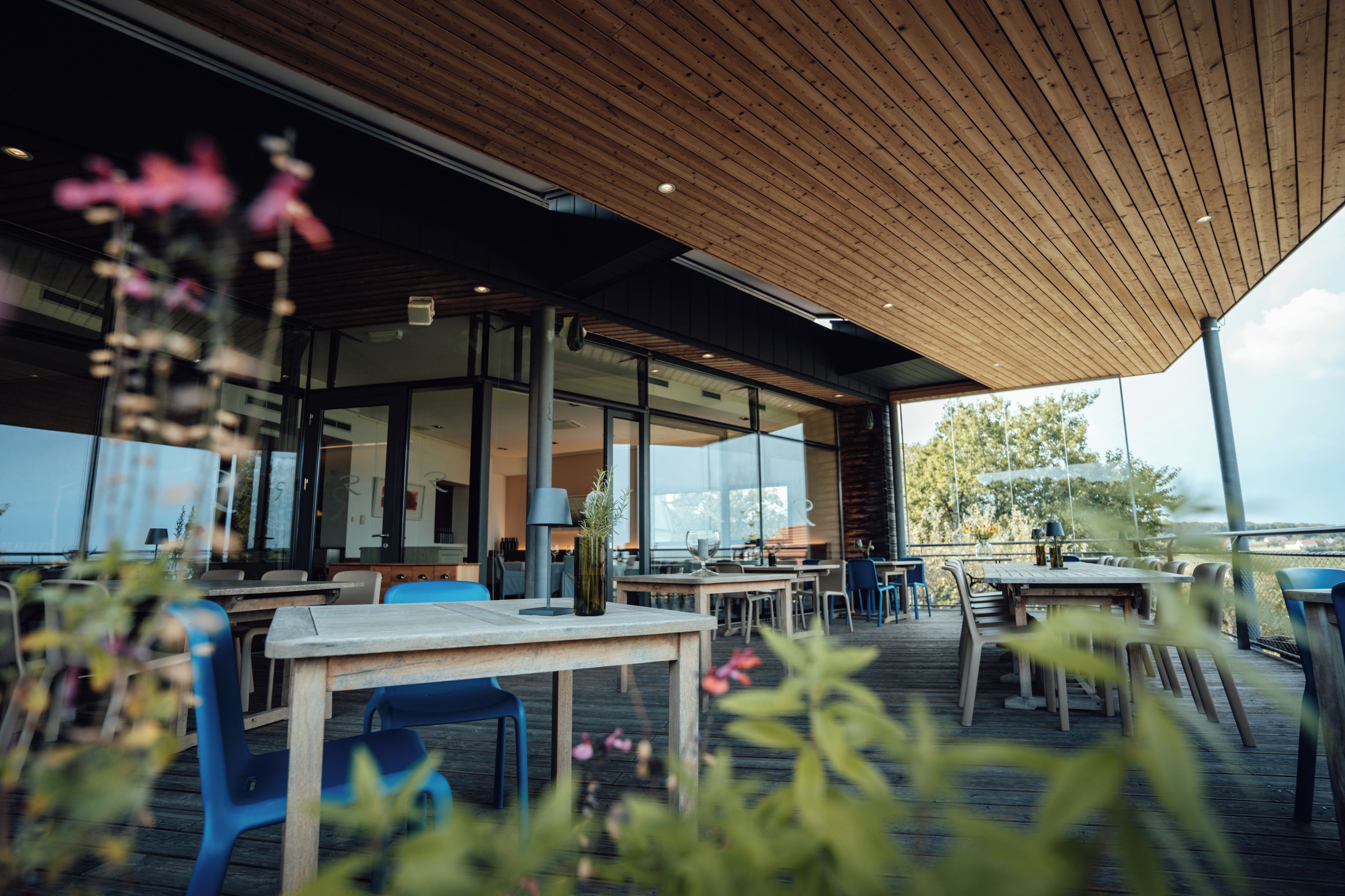 An outdoor seating area with wooden tables and chairs, a glass wall, and a view of the outdoors.